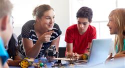 female teacher talking to four adolescent students while holding a small robot